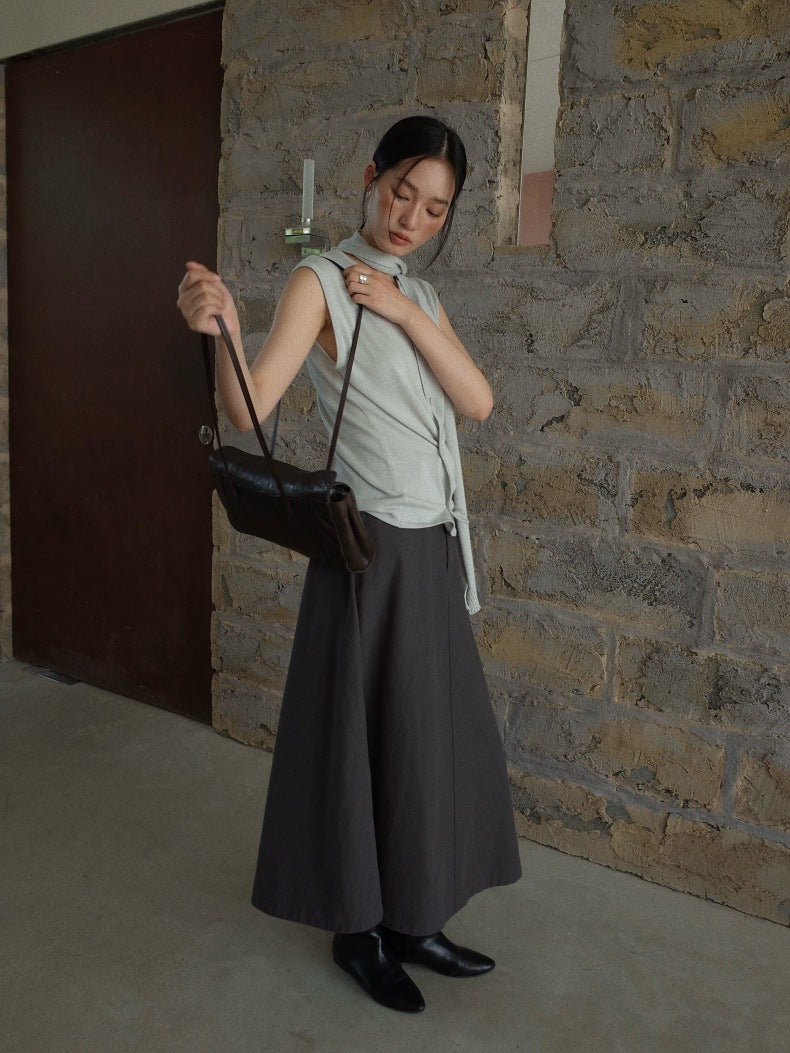 A woman stands in a rustic stone hallway wearing a light sage green sleeveless top, a dark gray wide-leg Maxi Length A-Line Denim Skirt, and black shoes, while holding a black leather shoulder bag.