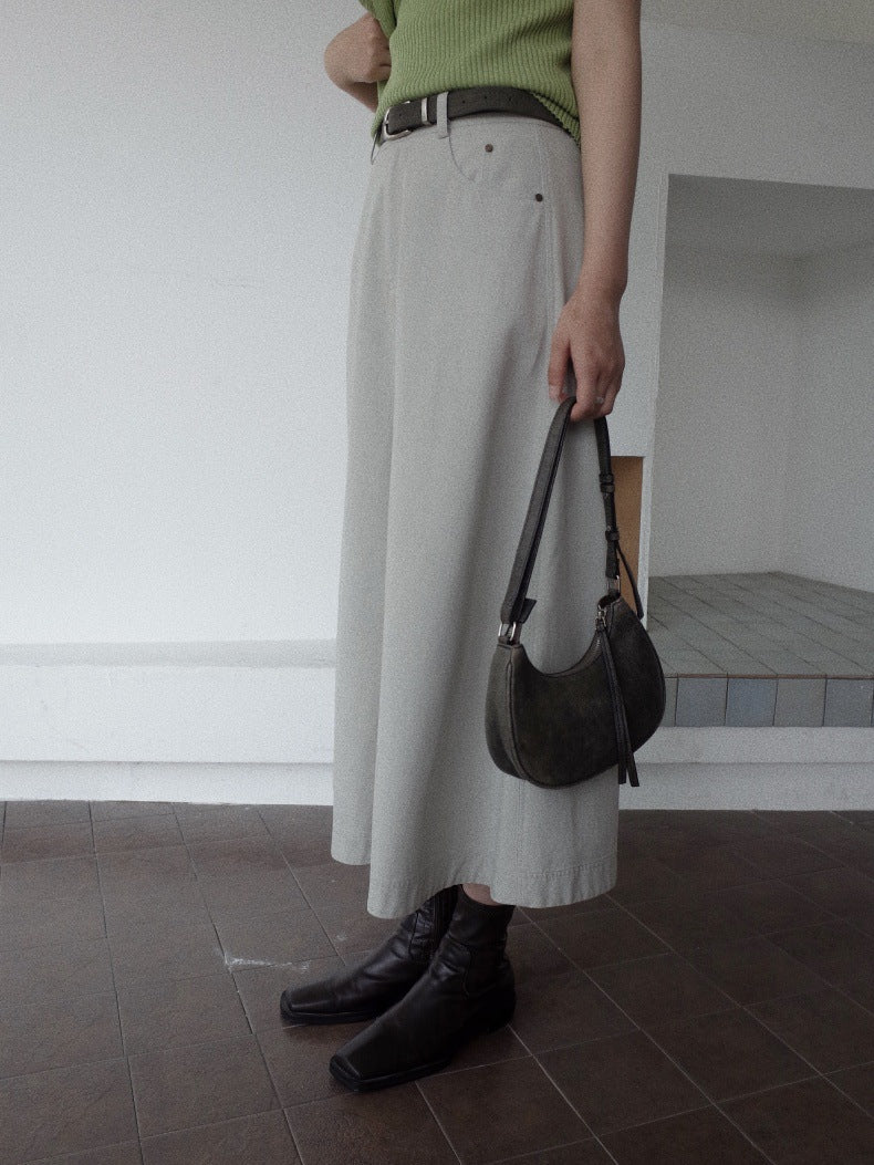 A person wearing a light beige Maxi Length A-Line Denim Skirt, a green ribbed top, black boots, and holding a dark brown crescent-shaped shoulder bag in an indoor setting.