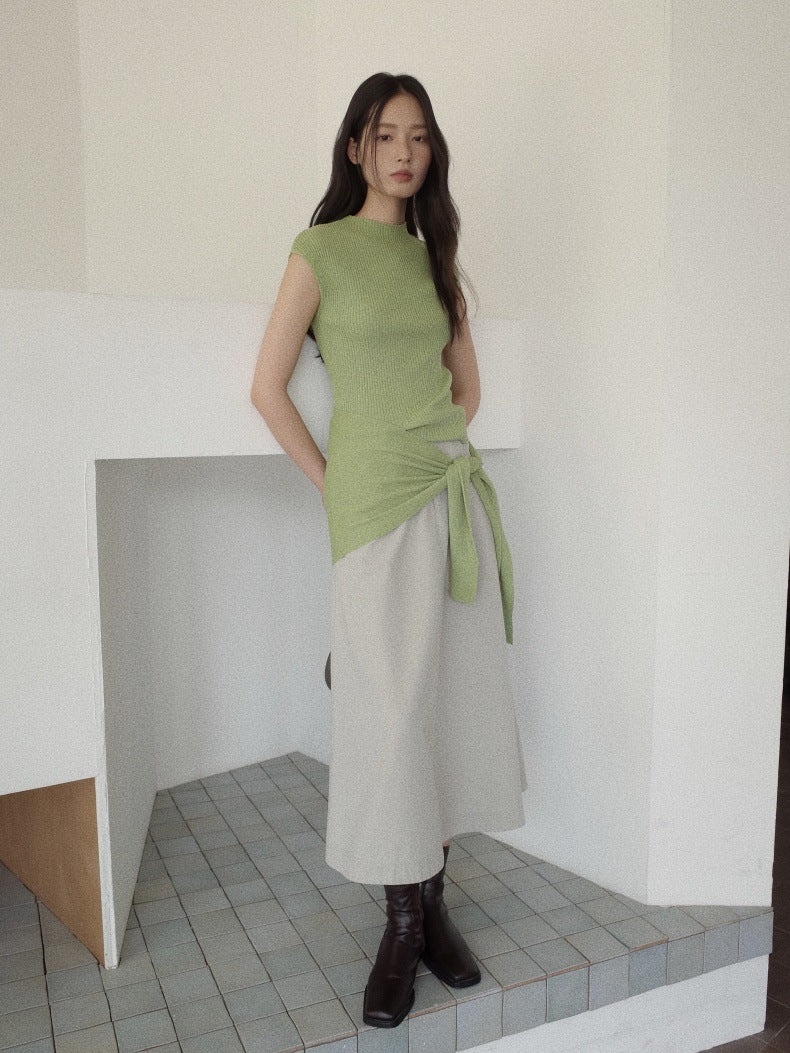 A woman models a light green sleeveless knit top with a side-tie detail, paired with a light gray Maxi Length A-Line Denim Skirt and dark brown ankle boots, standing in a minimalist white room with tiled flooring.