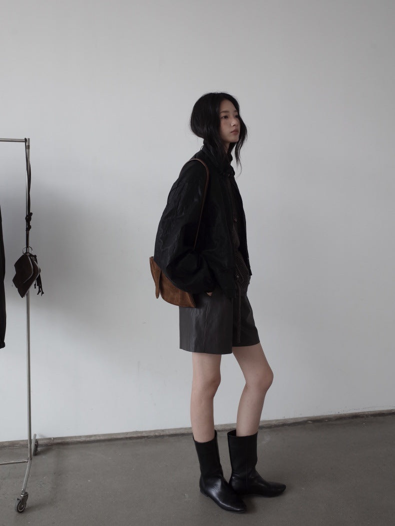 A woman stands in profile wearing a black jacket, dark brown leather shorts, black socks, and black boots, with a brown shoulder bag, set against a plain white wall.