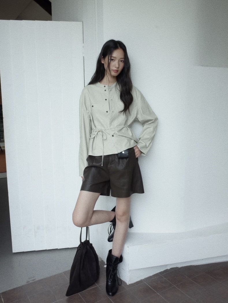 A woman posing in a light beige long-sleeve button-up shirt with a tie waist, paired with dark brown leather shorts, black ankle boots, and holding a black drawstring bag against a white wall.