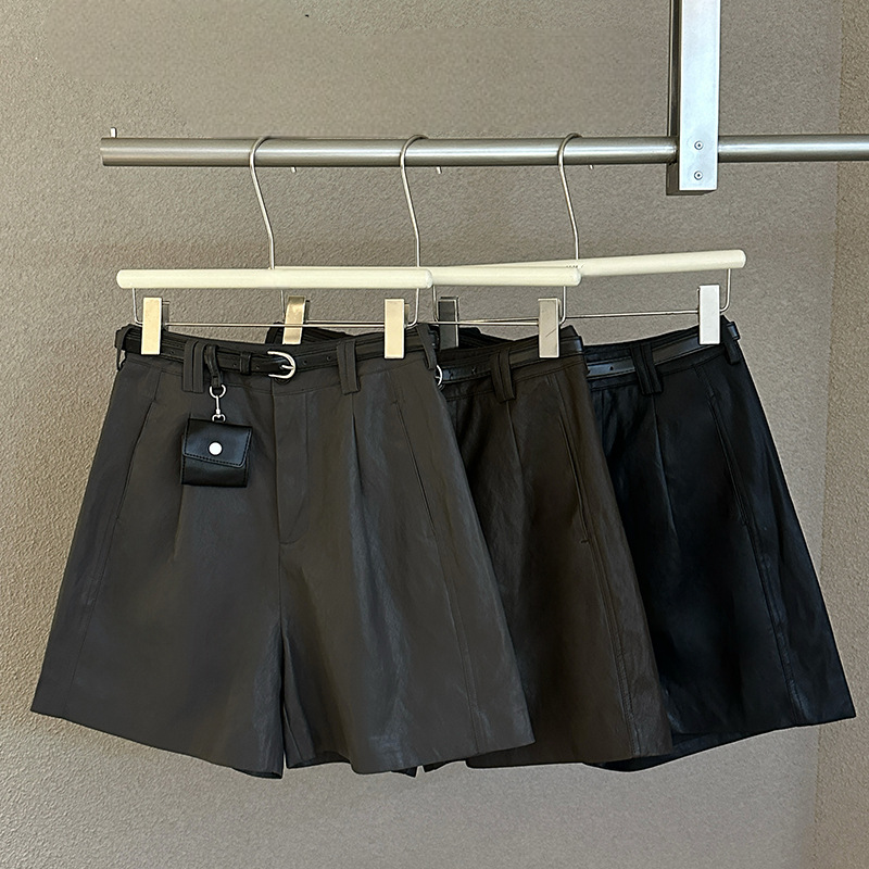 Three pairs of high-waisted shorts in black and dark brown leather fabric hang on white wooden hangers from a metal clothing rack against a textured beige wall.