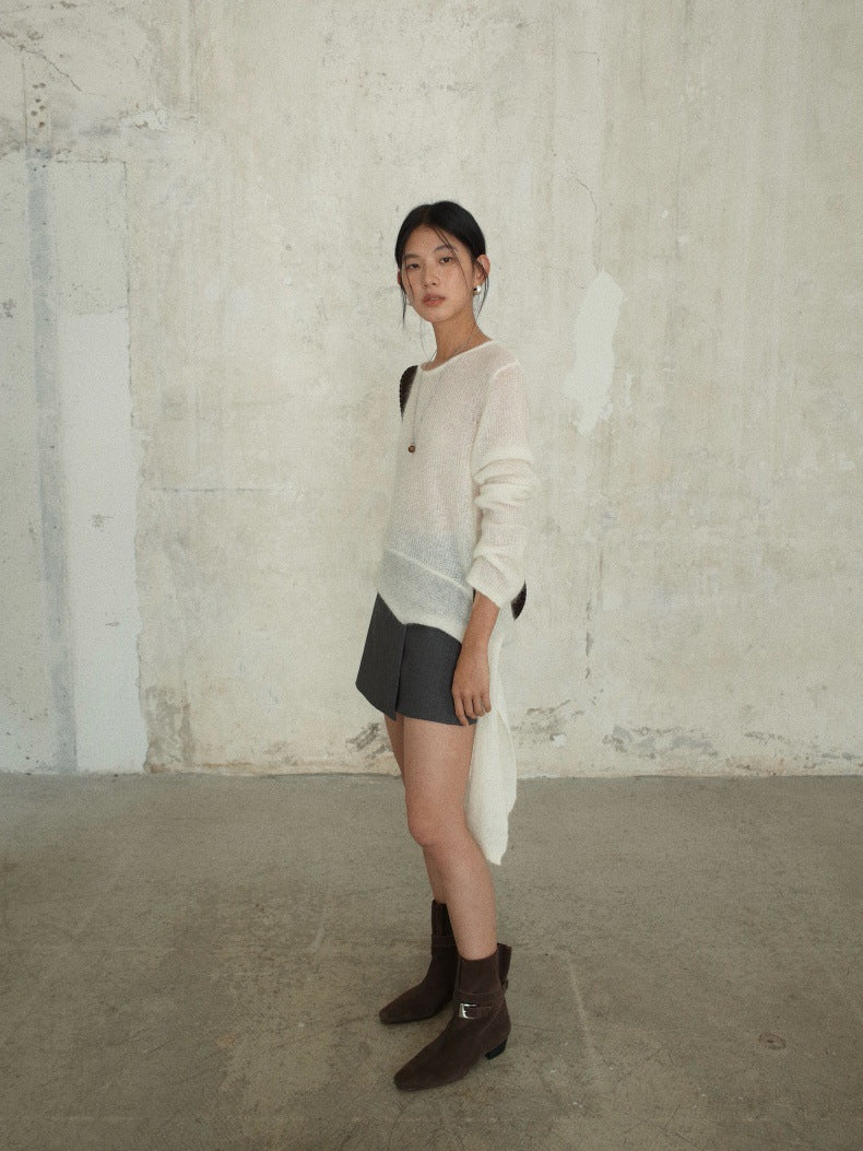 A woman stands in profile against a textured concrete wall, wearing a Strap Waist Long-sleeved Mohair Wool Knitted Cream Sweater with a dark gray mini skirt and dark brown ankle boots.