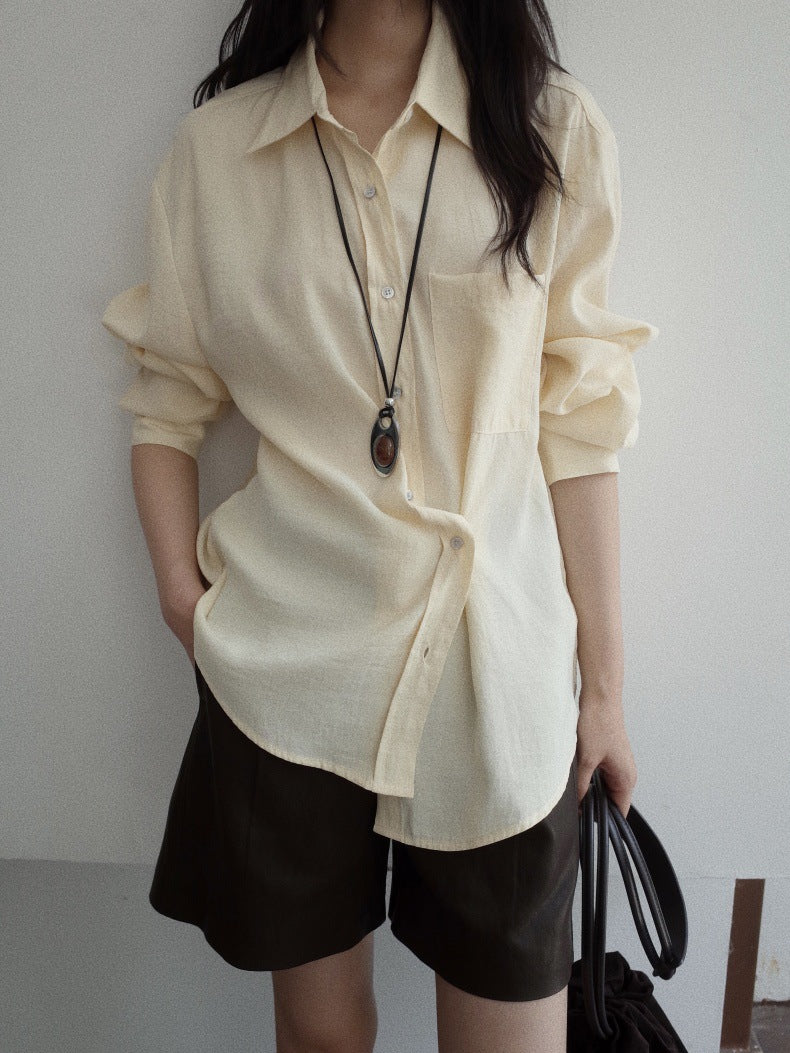 A woman wearing a cream-colored, long-sleeve button-down shirt and dark brown leather shorts, accessorized with a long black pendant necklace and holding a black handbag.