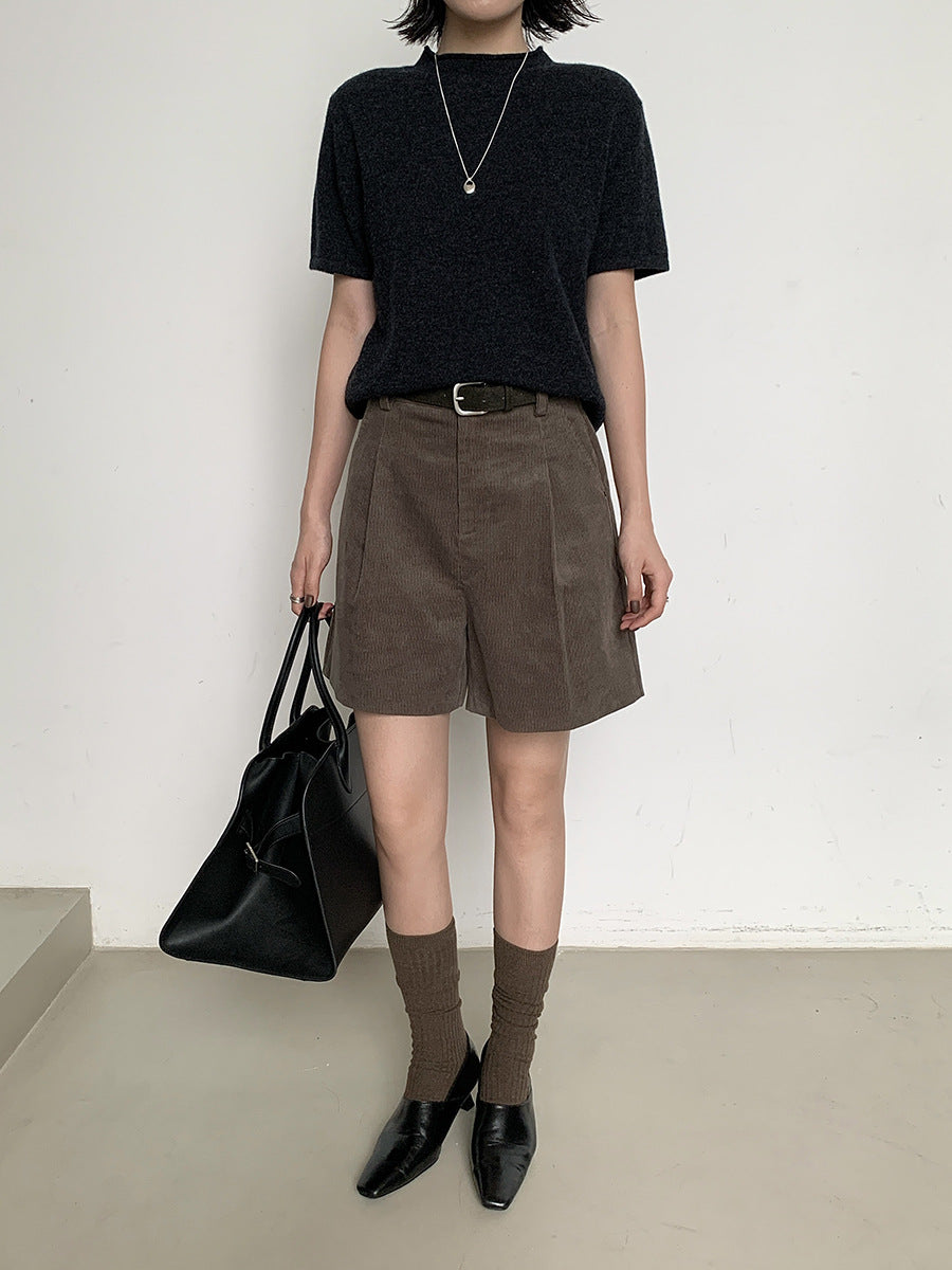Person wearing a black t-shirt, brown work dress Wide-leg shorts, and brown socks with black shoes, holding a black bag.