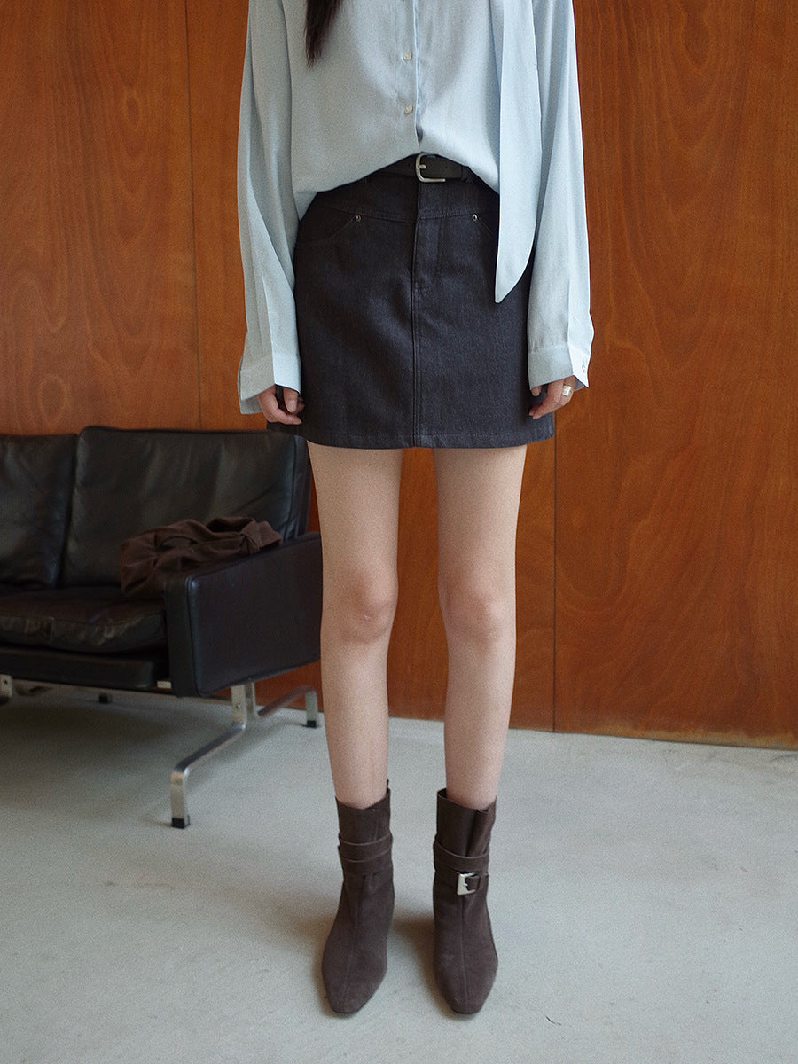 Woman wearing a white shirt, dark grey Denim High-waisted Short Skirt, and brown boots in a room with wooden walls and a black leather couch.