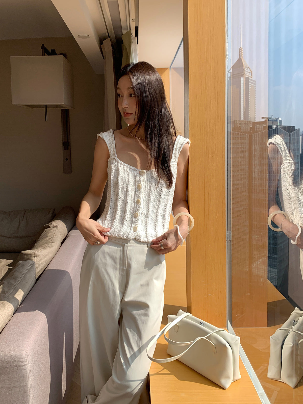 Woman in a white Thick Thread Knitted Cardigan Sleeveless Vest Short Tank Top and pants standing in a modern room with cityscape view.