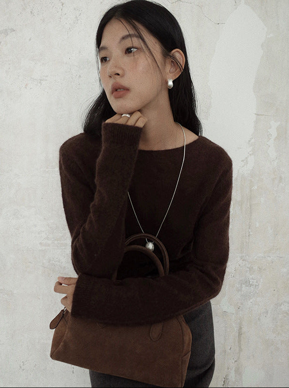 Woman wearing a brown wool pullover sweater holding a brown handbag against a light gray wall.