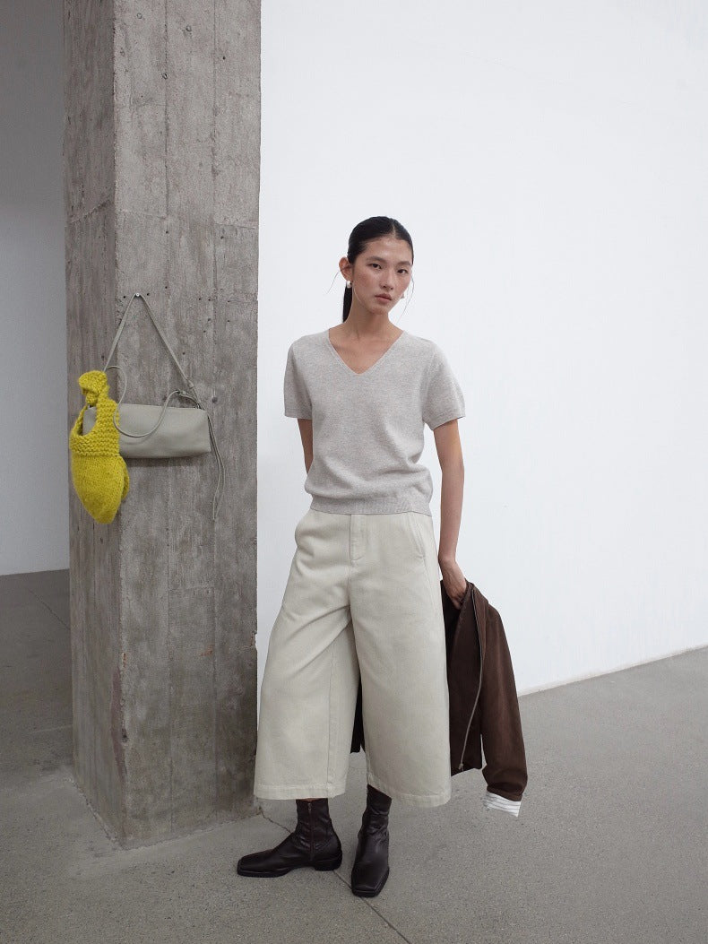 A model stands in a minimalist studio setting with a concrete pillar and white wall, wearing a light gray v-neck knit top, wide-leg cream jeans, and black boots, while holding a brown jacket.