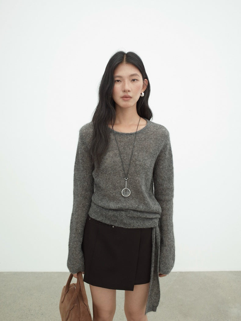 A woman stands against a plain light gray wall wearing a heather gray long-sleeve Strap Waist Long-sleeved Mohair Wool Knitted Sweater with a side tie detail, a black mini skirt, a long silver pendant necklace, and holding a brown leather tote bag.