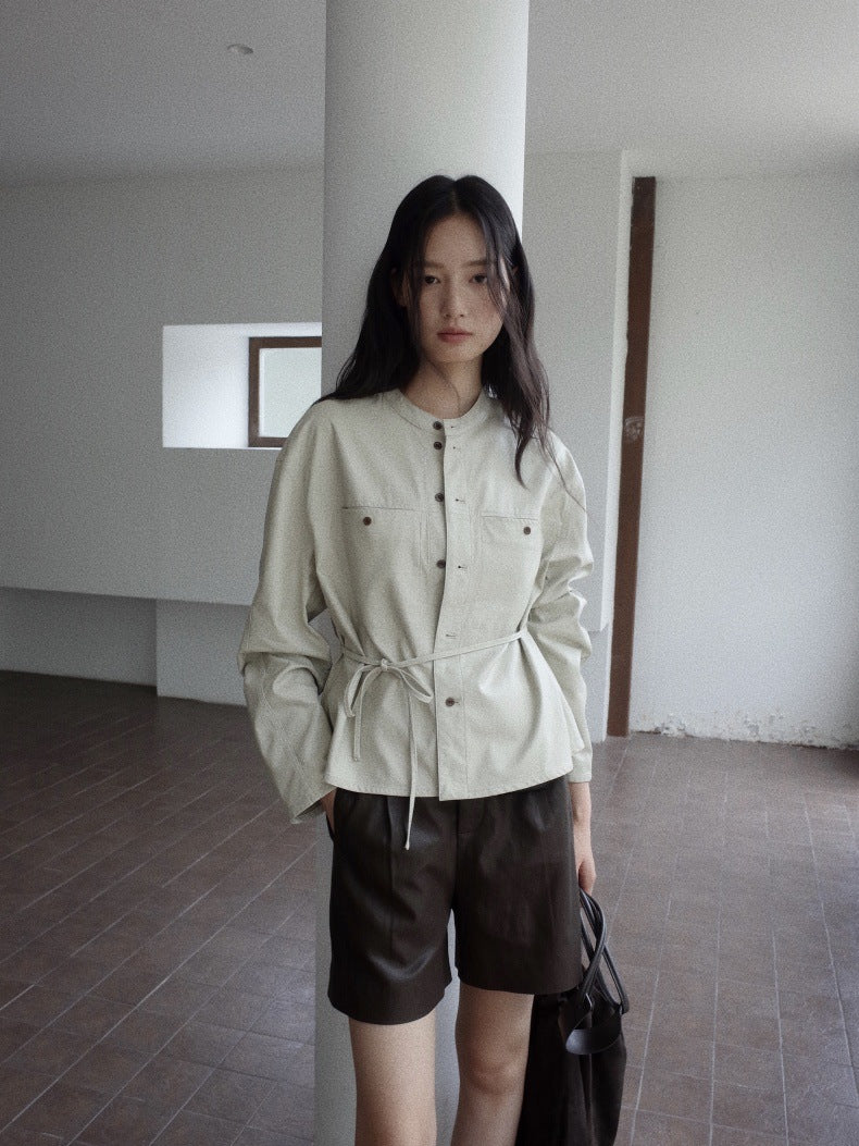 A woman stands indoors wearing a light beige cropped jacket with a drawstring waist and dark brown leather shorts, holding a black bag in a minimalist room with white walls and tiled flooring.