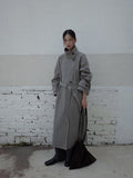 Person wearing a long gray Stand-up Collar Long Trench Coat with Belt standing against a white brick wall.
