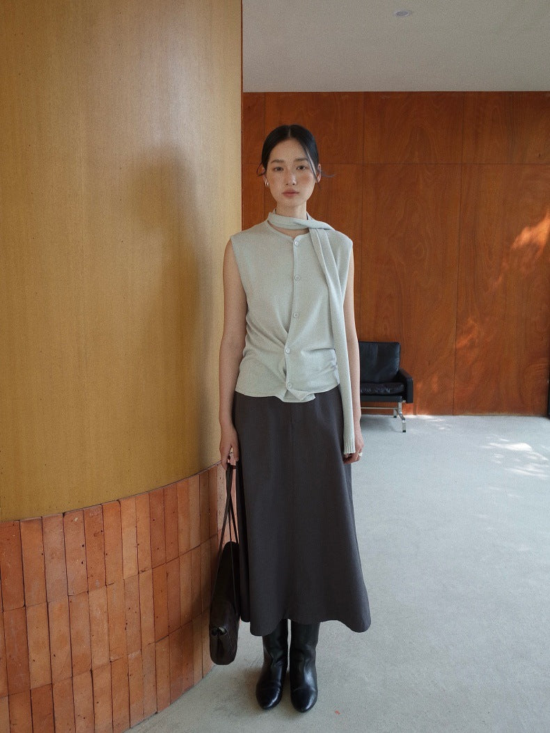 A woman stands indoors wearing a sleeveless light beige top with a high neck and front twist detail, paired with a dark charcoal Maxi Length A-Line Denim Skirt and black boots, holding a dark shoulder bag.