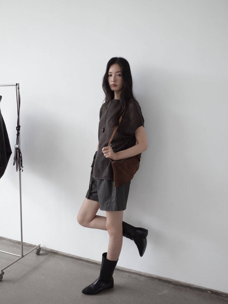 A woman stands against a white wall wearing a dark brown short-sleeve button-front top, dark gray leather shorts, black ankle boots, and carrying a brown shoulder bag.