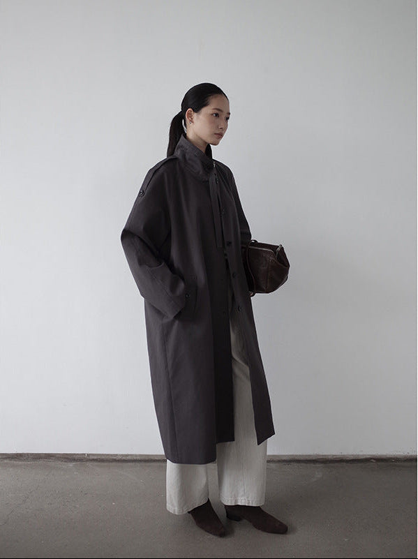 Woman wearing a dark Stand-up Collar Long Relaxed Fit Trench Coat and white pants against a plain background