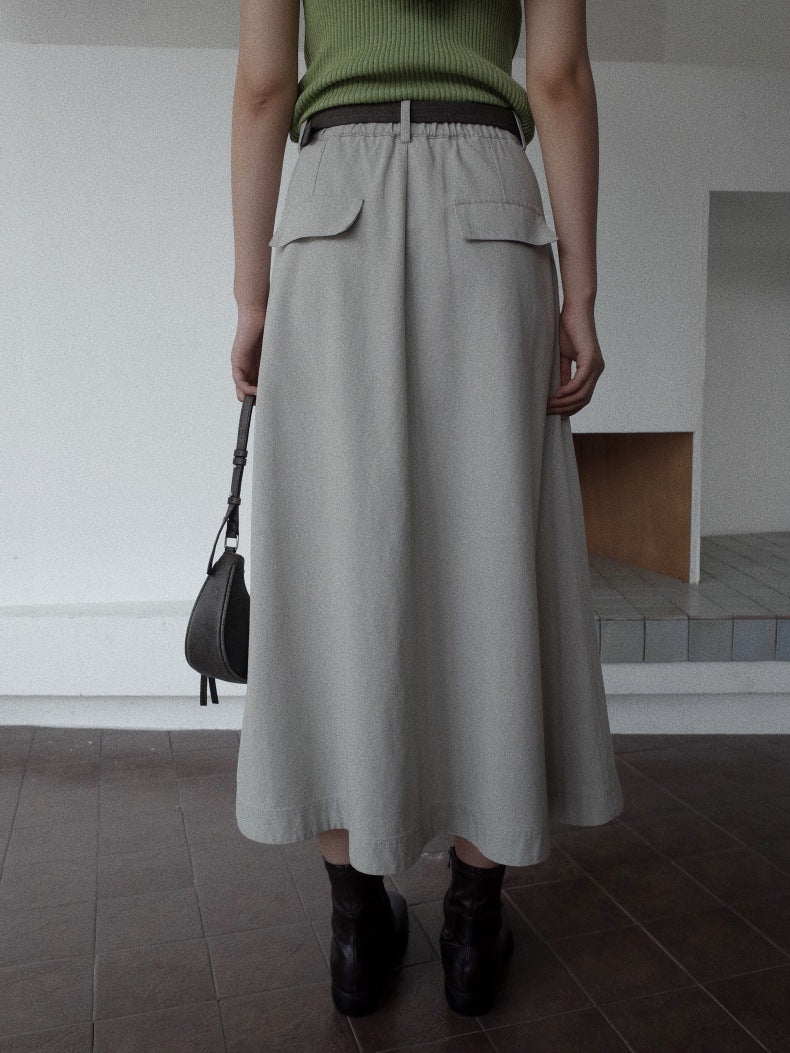 A person shown from the back wearing a light gray pleated Maxi Length A-Line Denim Skirt with flap pockets, paired with a dark belt, black ankle boots, and holding a dark gray handbag in an indoor minimalist setting.