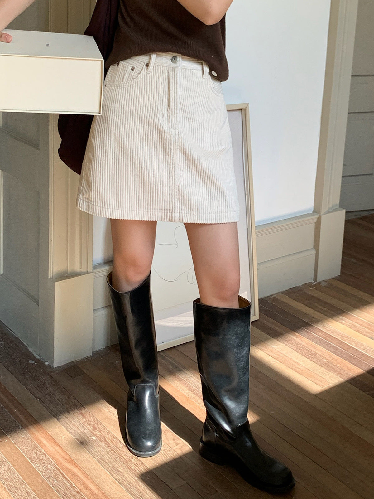 Person wearing a white striped Retro Corduroy A-line Short High-waisted mini skirt and black knee-high boots on a wooden floor.