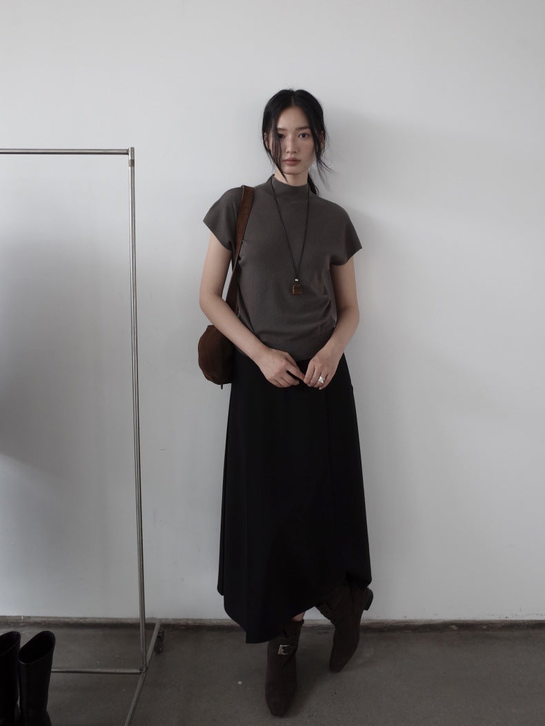 A woman stands indoors wearing a dark taupe short-sleeve mock-neck top, a long black Maxi Length Drape A-Line Skirt, and dark brown boots, with a brown shoulder bag. She is posed in front of a plain white wall next to a clothing rack.