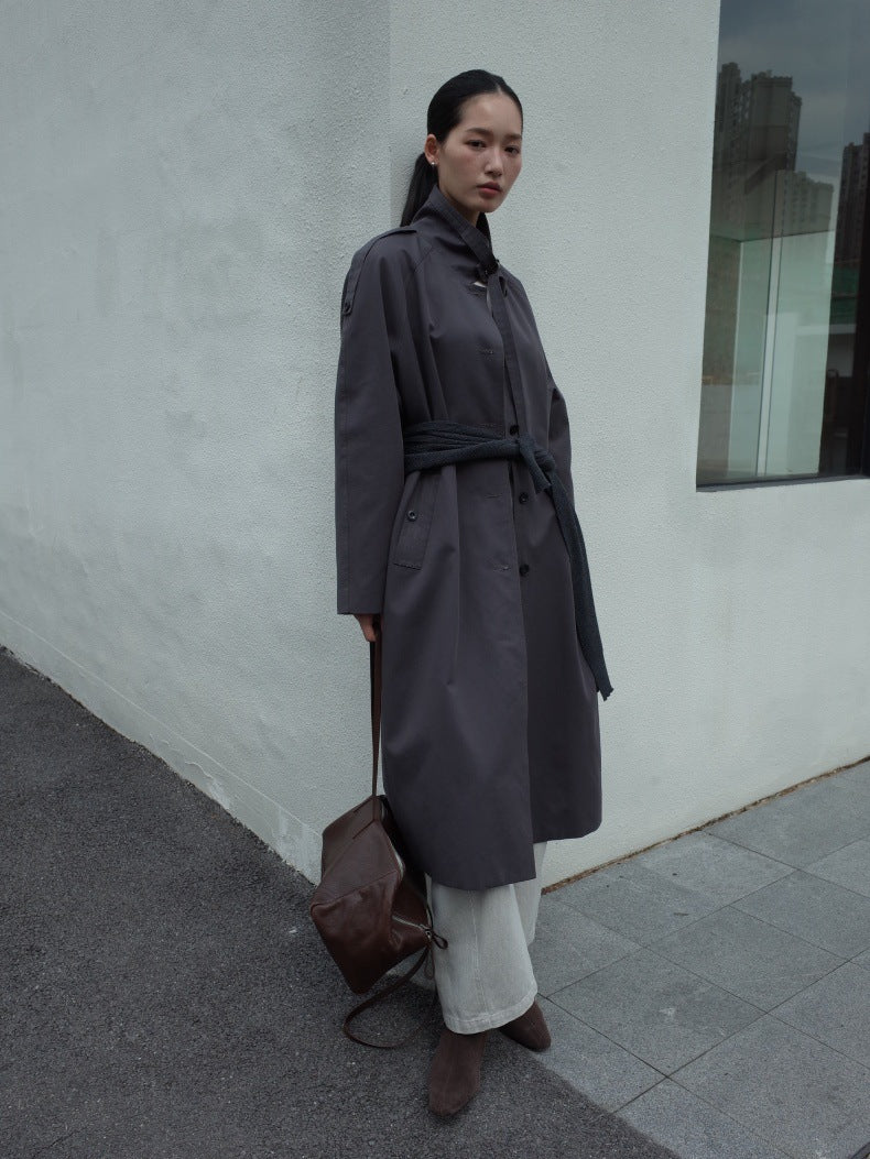 A woman stands outdoors against a white wall wearing a long dark gray belted trench coat over a black top and light gray wide-leg pants, with brown ankle boots and a brown shoulder bag.