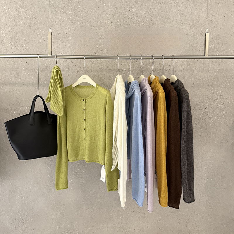 A curated display of knit apparel hanging on a metal rack against a textured gray wall, including a black tote bag, a light olive green cardigan, a cream top, a light blue top, a lavender top, a mustard yellow top, a brown top, and a dark gray top.
