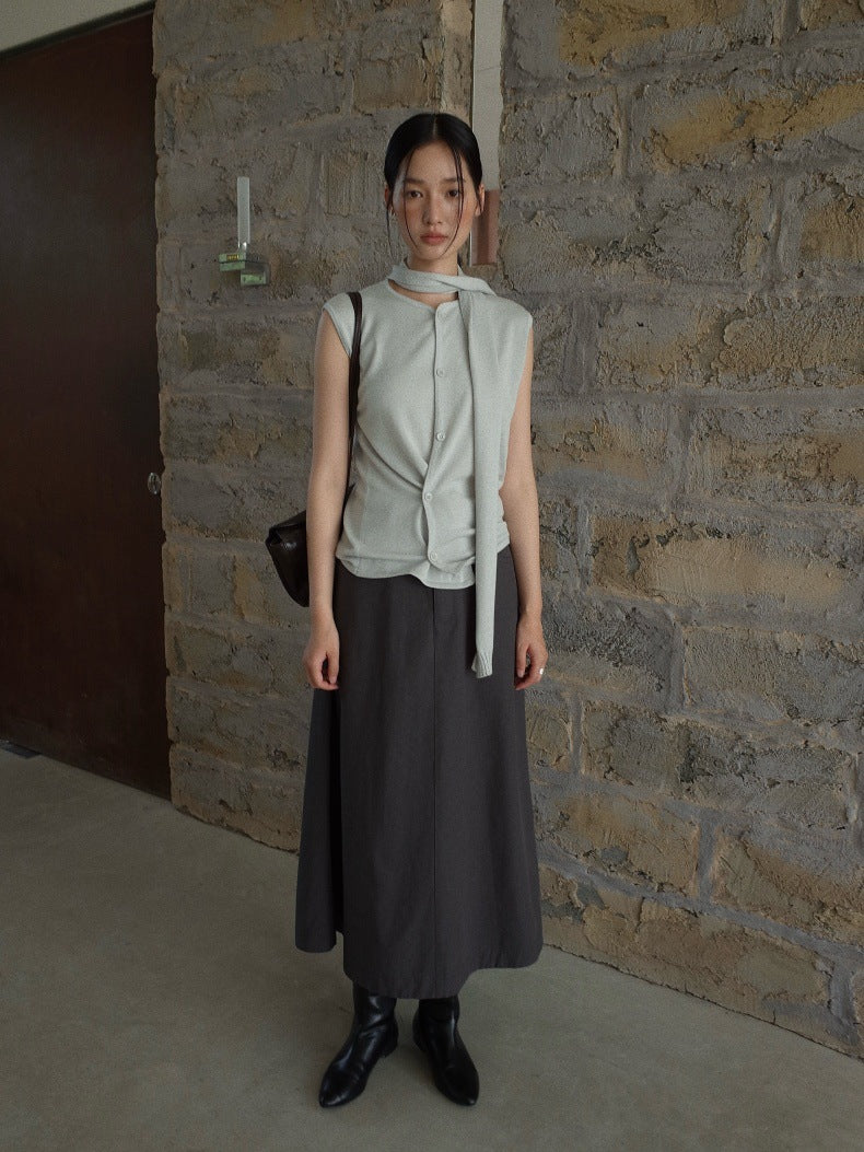 A woman stands against a rustic stone wall wearing a light sage green sleeveless top with a front tie detail and a dark charcoal gray midi Maxi Length A-Line Denim Skirt, paired with black ankle boots and a small black shoulder bag.