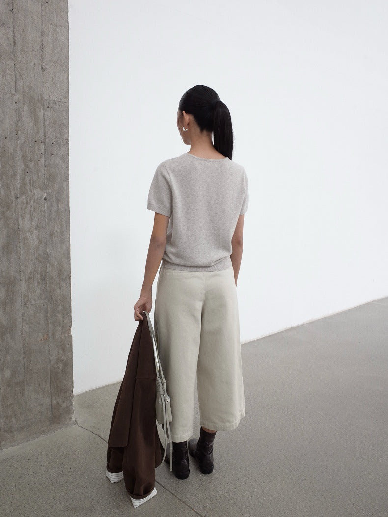 A woman stands with her back to the camera in a minimalist studio setting, wearing a light gray short-sleeve knit top, cream wide-leg cropped jeans, and black ankle boots. She is holding a dark brown coat and a white bag.