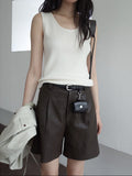 Person wearing a white sleeveless top and brown leather shorts with a small black bag, standing against a plain background.