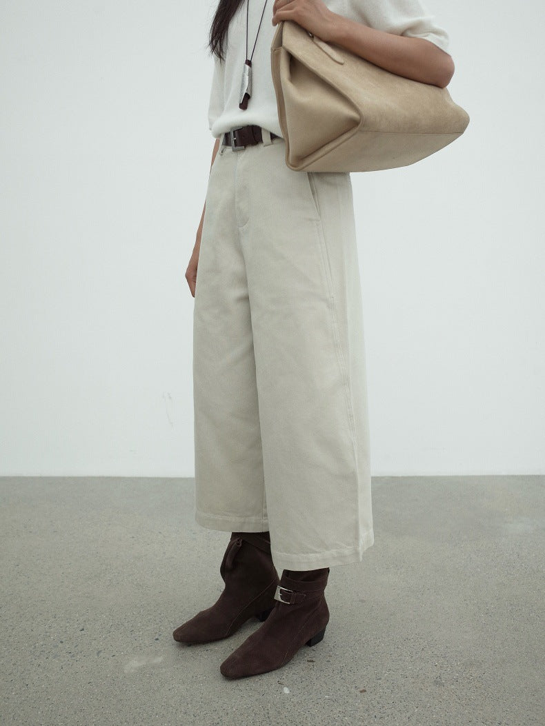 A person wearing wide-leg cropped jeans in a light beige color, paired with dark brown ankle boots and holding a large beige shoulder bag, set against a minimalist studio background.