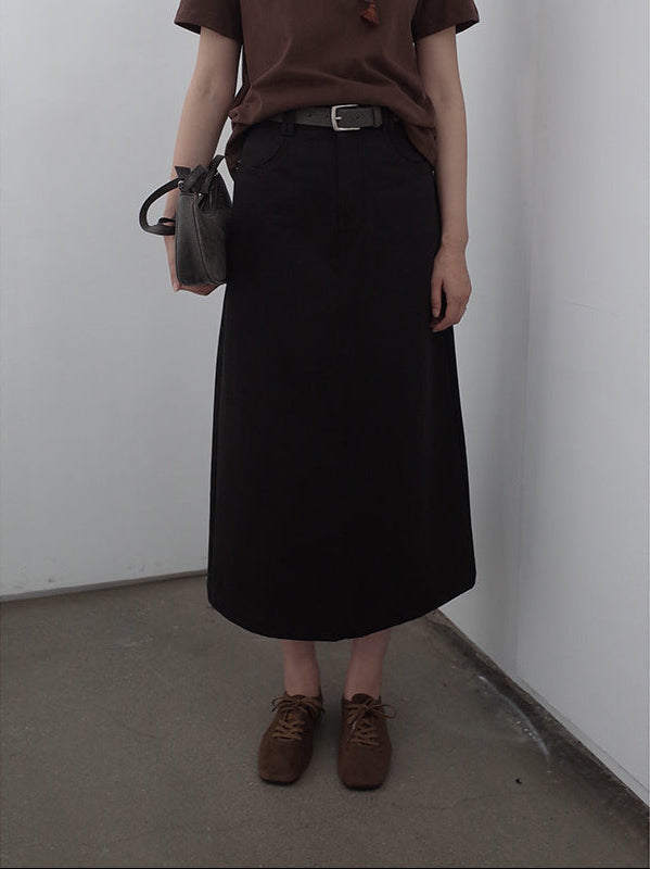 Person wearing a brown shirt, black A-line Midi Denim Skirt, and brown shoes against a plain wall.
