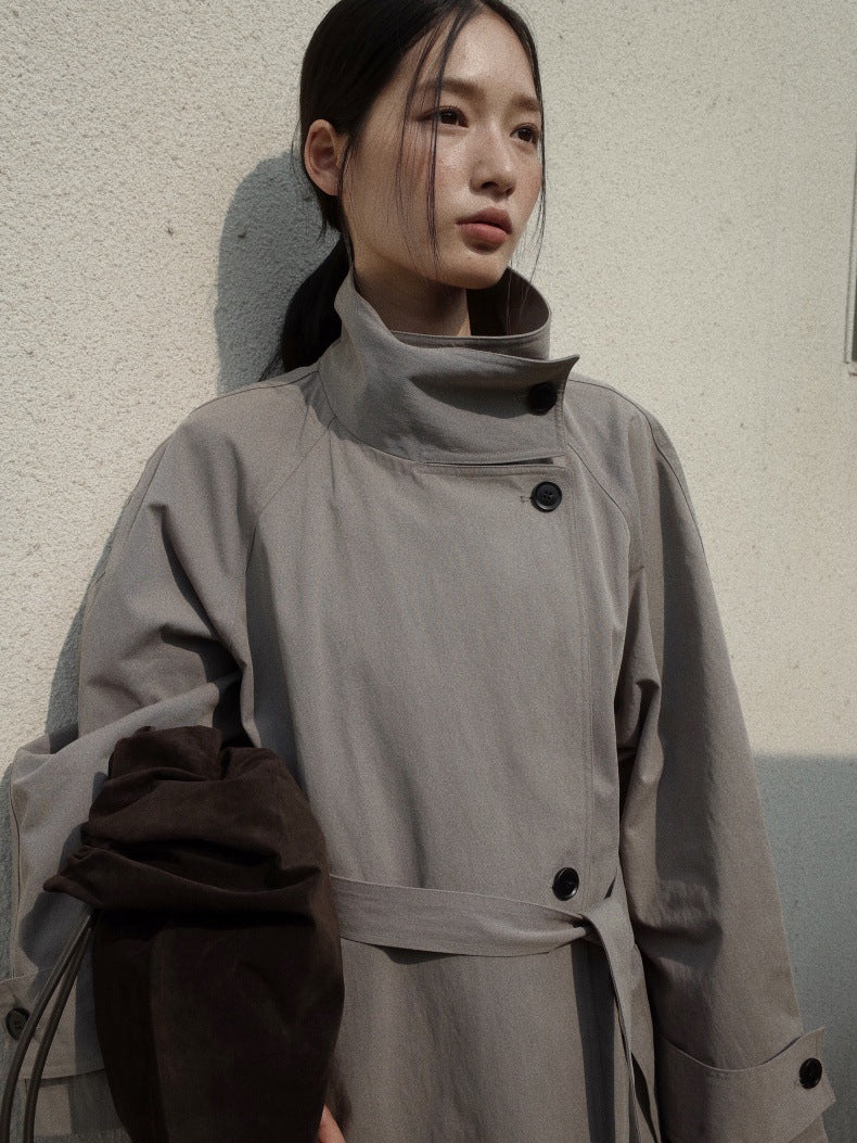 A woman wearing a light gray wrap-style trench coat with a high collar and dark buttons, leaning against a textured off-white wall.