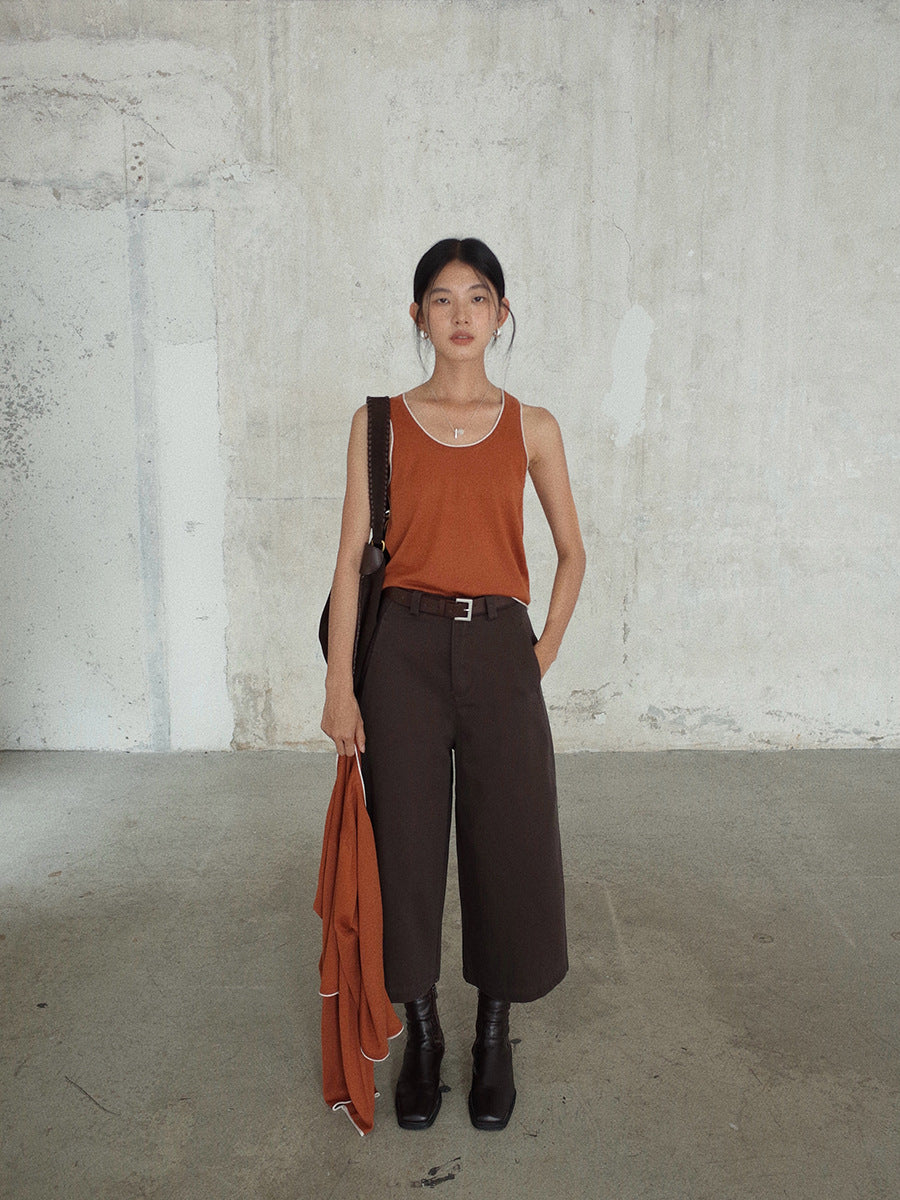 Woman wearing a rust-colored tank top and dark Cropped Wide-Leg Straight Jeans, holding an orange garment against a concrete wall.