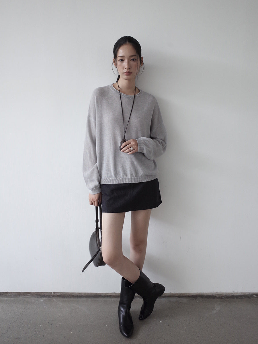 Woman wearing a gray wool sweater, black skirt, and black boots against a white wall.