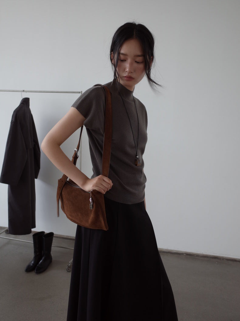 A woman wearing a dark gray mock-neck short-sleeve top and a long black Maxi Length Drape A-Line Skirt, carrying a brown suede shoulder bag with a brown leather strap in a minimalist room.