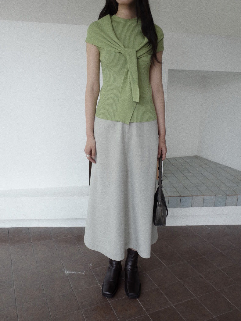 A woman wearing a light green short-sleeve knit top with a tied neckline and a light Maxi Length A-Line Denim Skirt, paired with black ankle boots and holding a black handbag, standing in a minimalist room with white walls and a tiled floor.