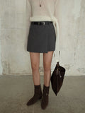 Person wearing a gray Irregular A-Line Short Mini Skirt, beige sweater, and brown boots against a textured wall.