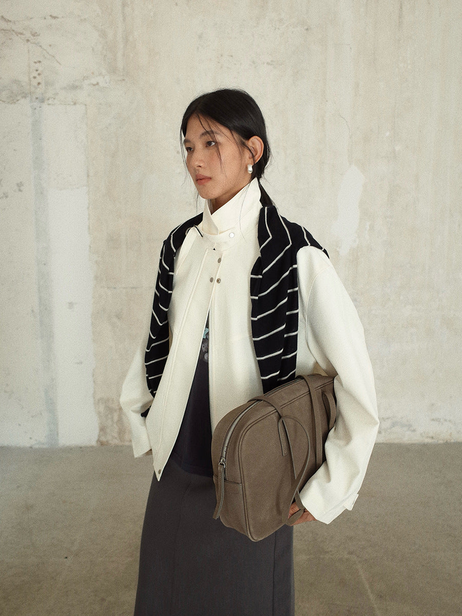 Person wearing a white Stand-up Collar Jacket , black and white striped vest, and holding a brown bag against a textured wall.
