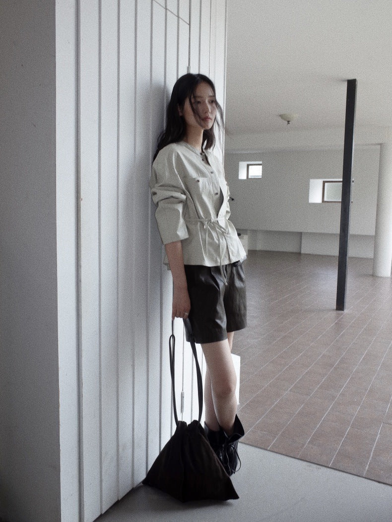 A woman stands in a minimalist indoor space with white paneled walls, wearing a light beige long-sleeve shirt, black leather shorts, and black boots, while holding a black tote bag.