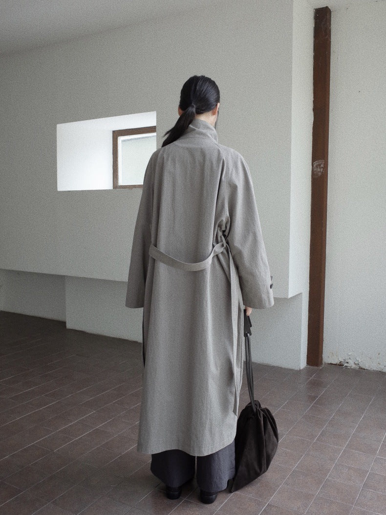 A person viewed from behind wearing a long light gray belted coat, standing in a minimalist room with white walls and a tiled floor, holding a dark bag.