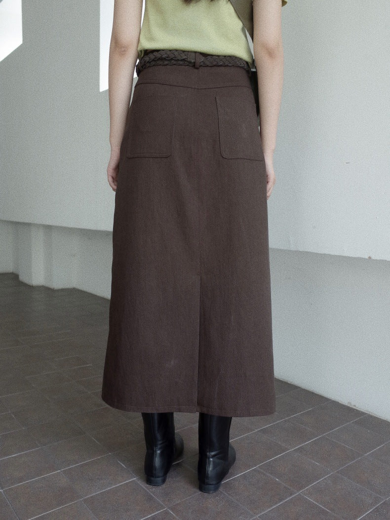 A person shown from the back wearing a dark brown long High-waisted A-line Maxi Skirt  skirt with two large patch pockets, a braided waistband, a light green top, and black ankle boots in an indoor setting with tiled floors and a white wall.