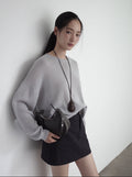 Woman in gray wool sweater and black skirt holding a black handbag against a white wall