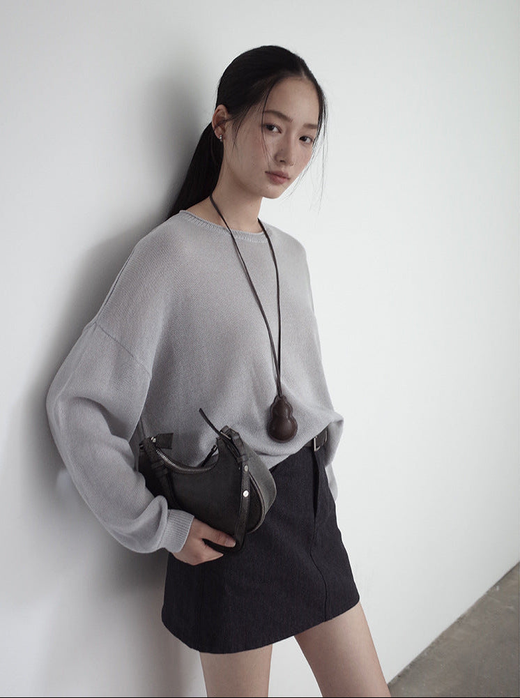 Woman in gray wool sweater and black skirt holding a black handbag against a white wall