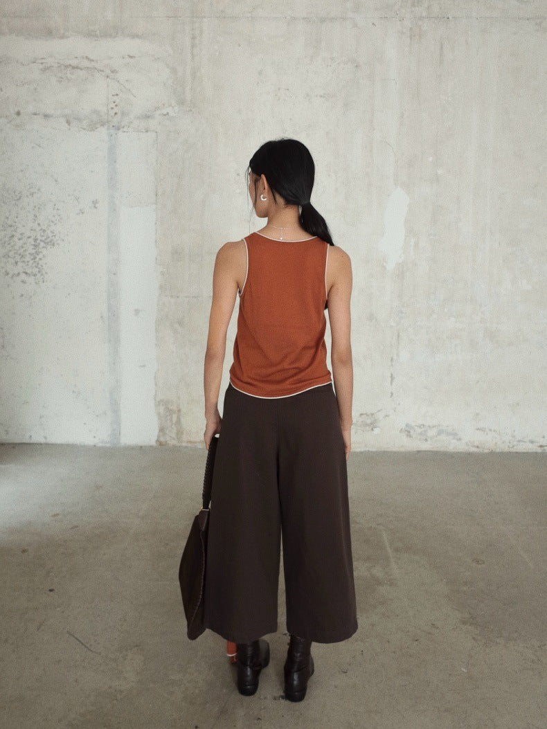 A woman stands with her back to the camera in a concrete room, wearing a rust-orange sleeveless top, dark brown Cropped Wide-Leg Straight Jeans, and black ankle boots, while holding a dark brown shoulder bag.