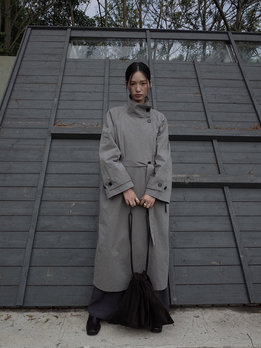 Person wearing a long gray Stand-up Collar Long Trench Coat with Belt standing in front of a wooden wall.