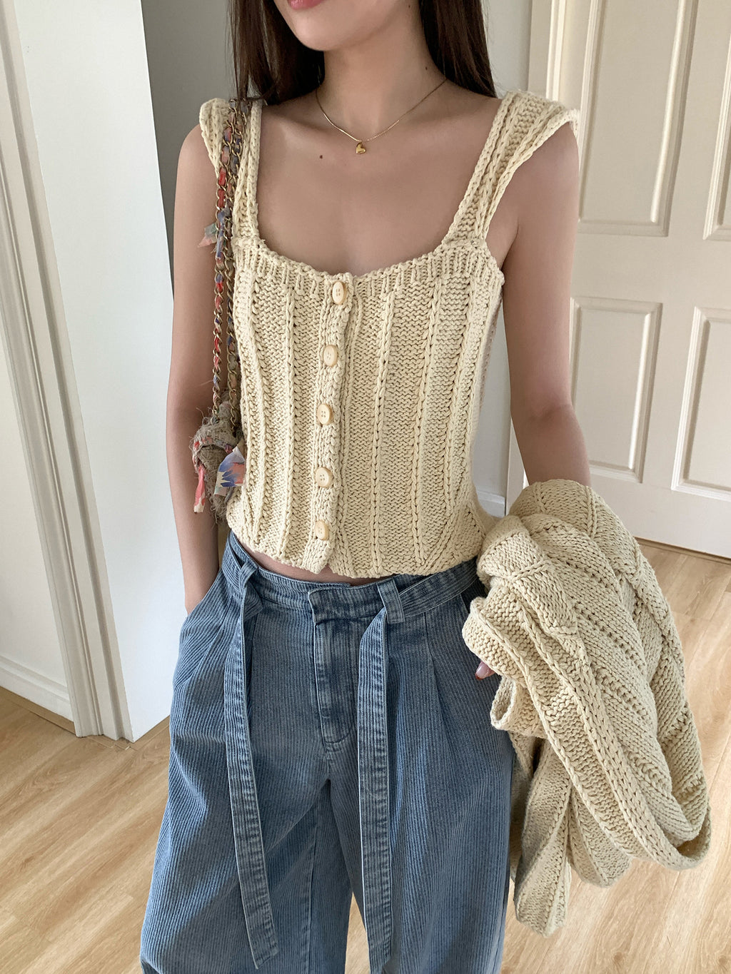Woman wearing a beige knitted tank top and blue jeans, holding a cardigan.