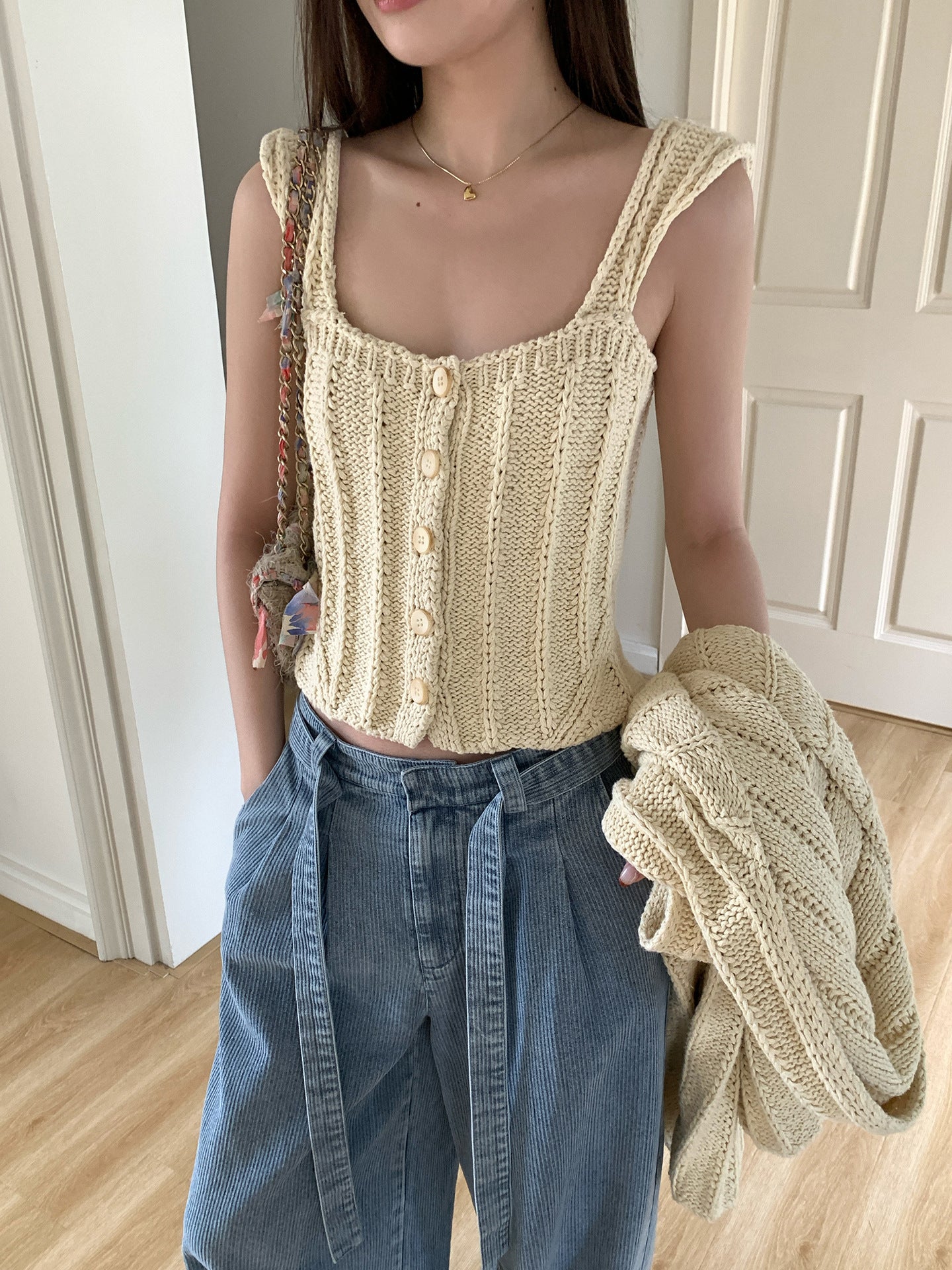 Woman wearing a beige knitted tank top and blue jeans, holding a cardigan.