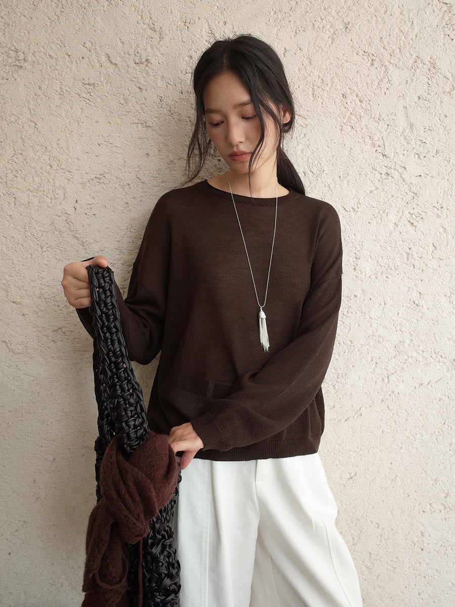 Woman wearing a brown wool sweater and white pants holding a dark scarf against a beige wall.