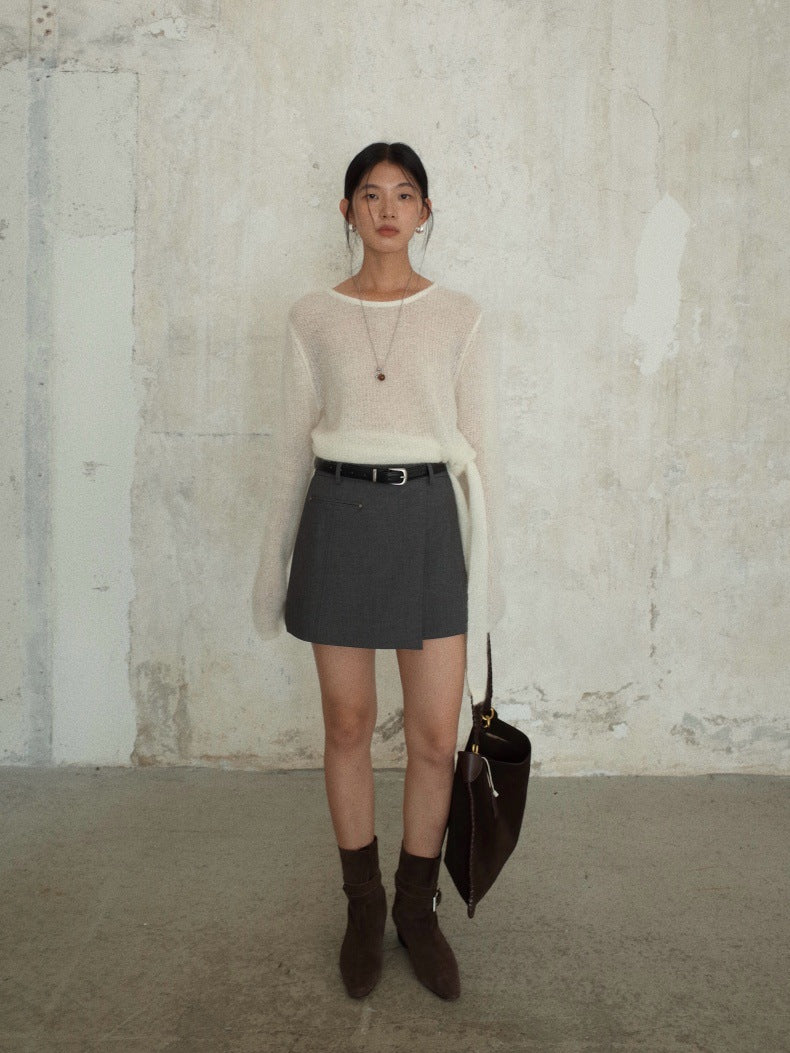 A woman stands against a textured concrete wall wearing a cream-colored sheer long-sleeve sweater layered over a white undershirt, paired with a dark gray mini skirt, a black belt, and dark brown ankle boots. She is holding a dark brown leather handbag.