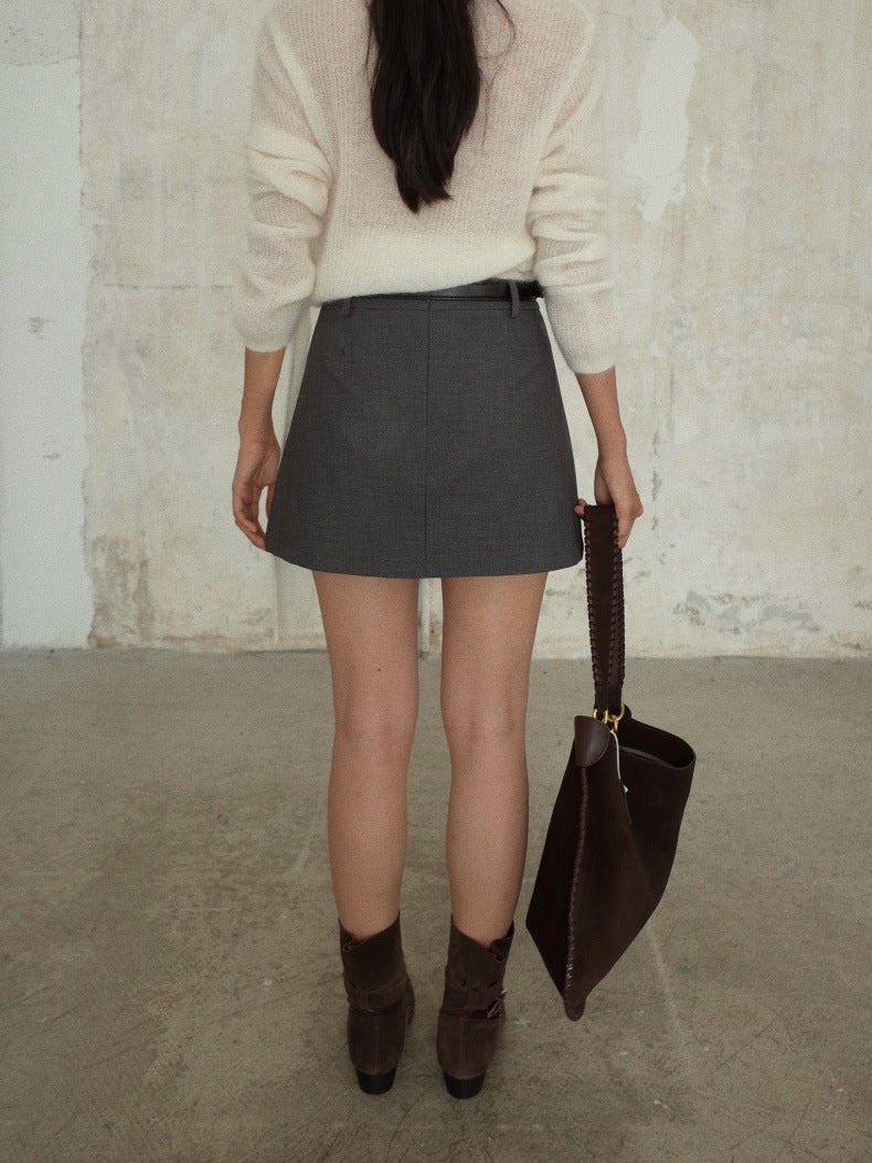A person shown from the back wearing a cream knit sweater, a dark gray Irregular A-Line Short Mini Skirt with a thin black belt, and dark brown ankle boots, while holding a dark brown leather shoulder bag against a textured concrete wall.