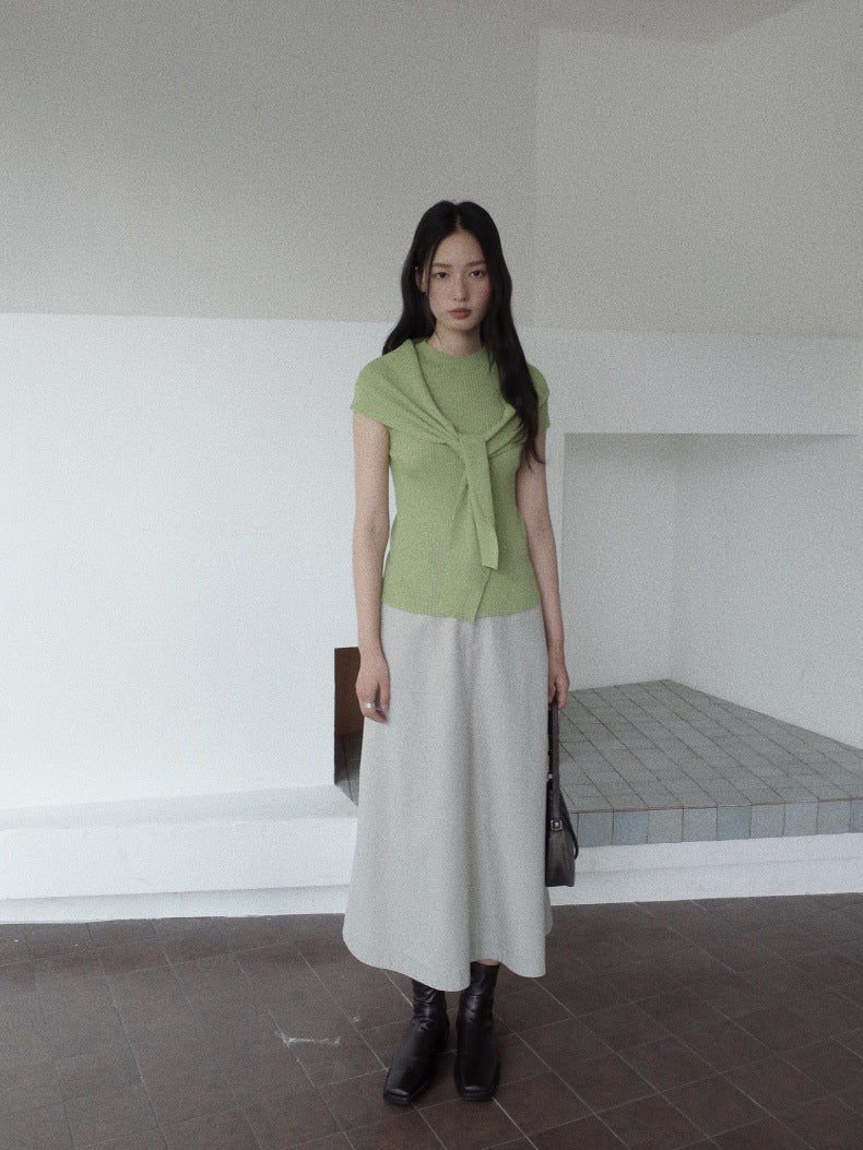 A woman stands in a minimalist white room wearing a light green short-sleeve knit top with a front tie detail, a long light gray Maxi Length A-Line Denim Skirt, and black ankle boots.