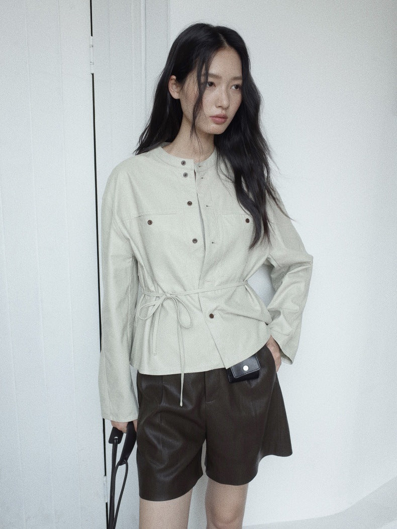 A woman modeling a light beige long-sleeve button-front jacket with a tie waist, paired with dark brown leather shorts, standing against a white wall.